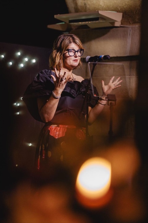 Lucy Littlewood speaking at a microphone, mid-gesture, lit by candlelight.